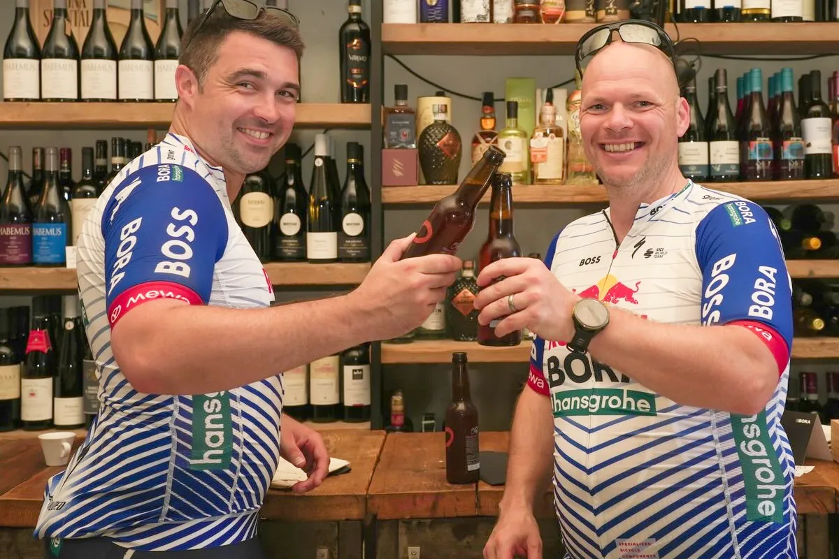 Due ospiti in maglia da ciclismo brindano davanti alla parete dei vini di MASTO